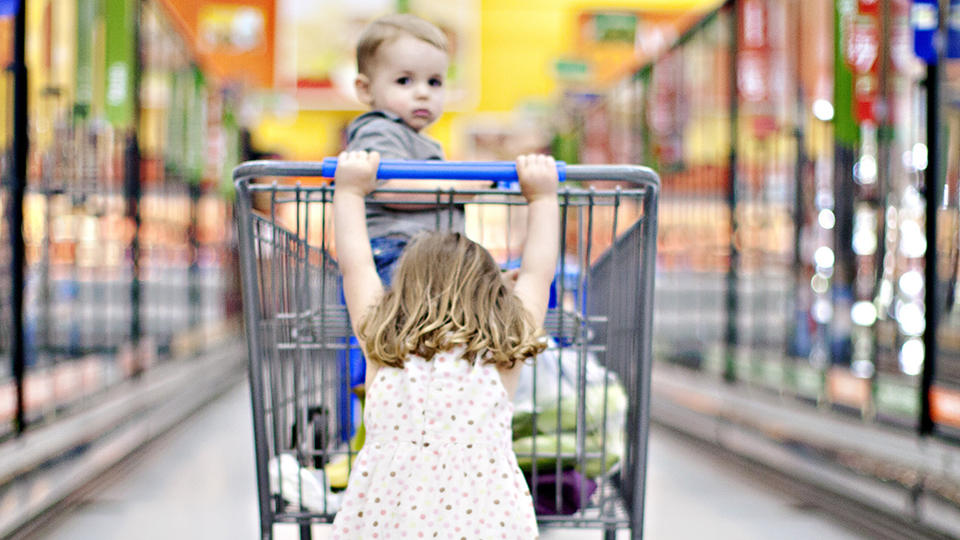 Little girl pushing younger sibling in a grocery cart. 